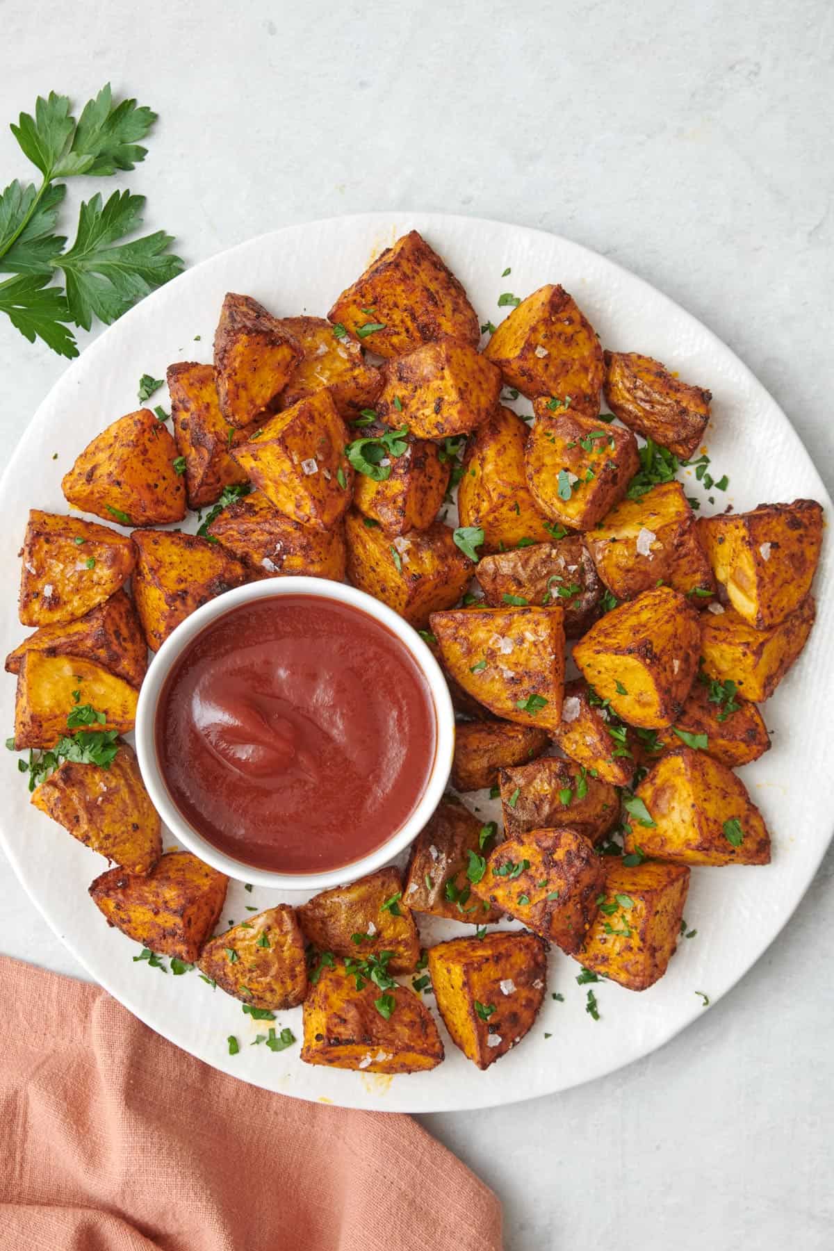 Round plate of crispy air fried seasoned potatoes garnished with flaky salt and fresh herbs, served with a small bowl of ketchup.