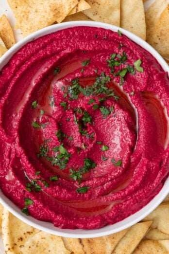 Beet hummus in a bowl garnished with fresh parsley that is set on a plate and surrounded with pita chips.