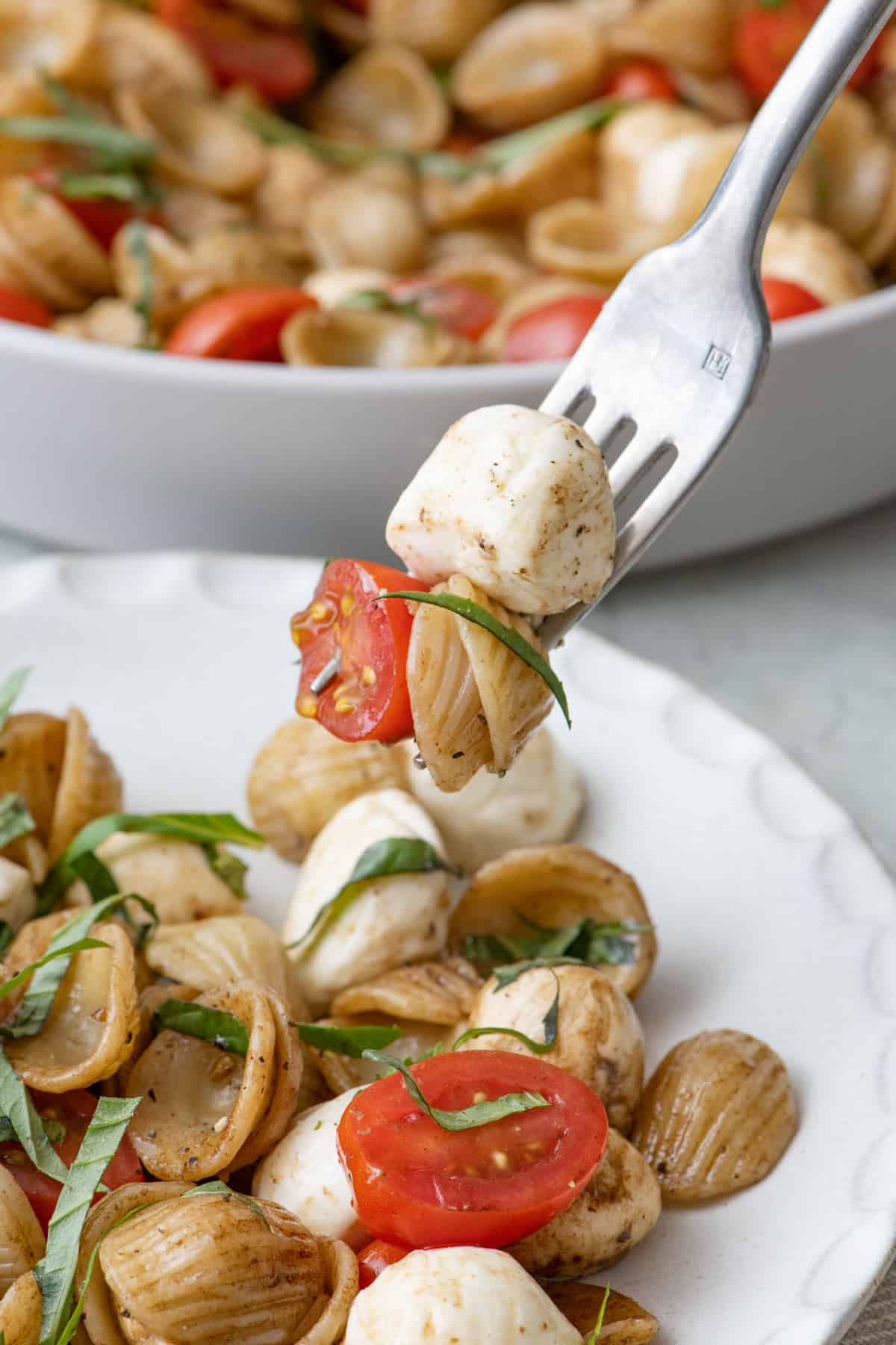 Fork lifting up a bite of caprese pasta salad with pasta, tomato, and cheese on it over a plate of the recipe and the serving bowl nearby.