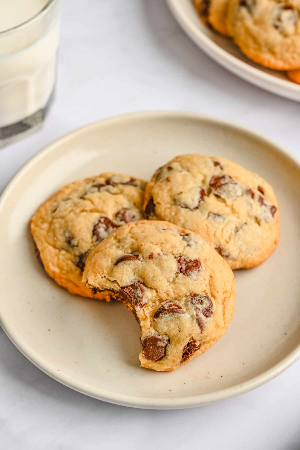 2 chocolate chip cookies on a smaller round plate with a bite taken out of one, full plate in the background with milk nearby.