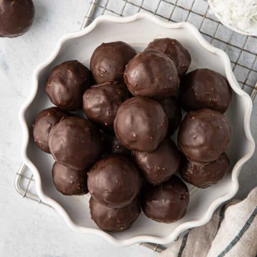 Chocolate coconut balls on a decorative plate with extra shredded coconut and melted chocolate nearby.