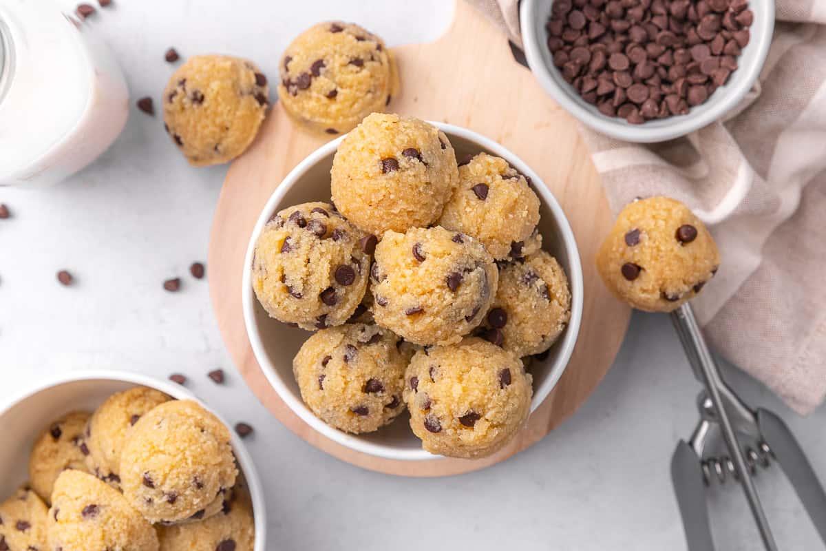 Two small bowls of edible cookie dough balls with one in the center. Cookie scoop with come dough on it, a few extra dough balls and chocolate chips around.