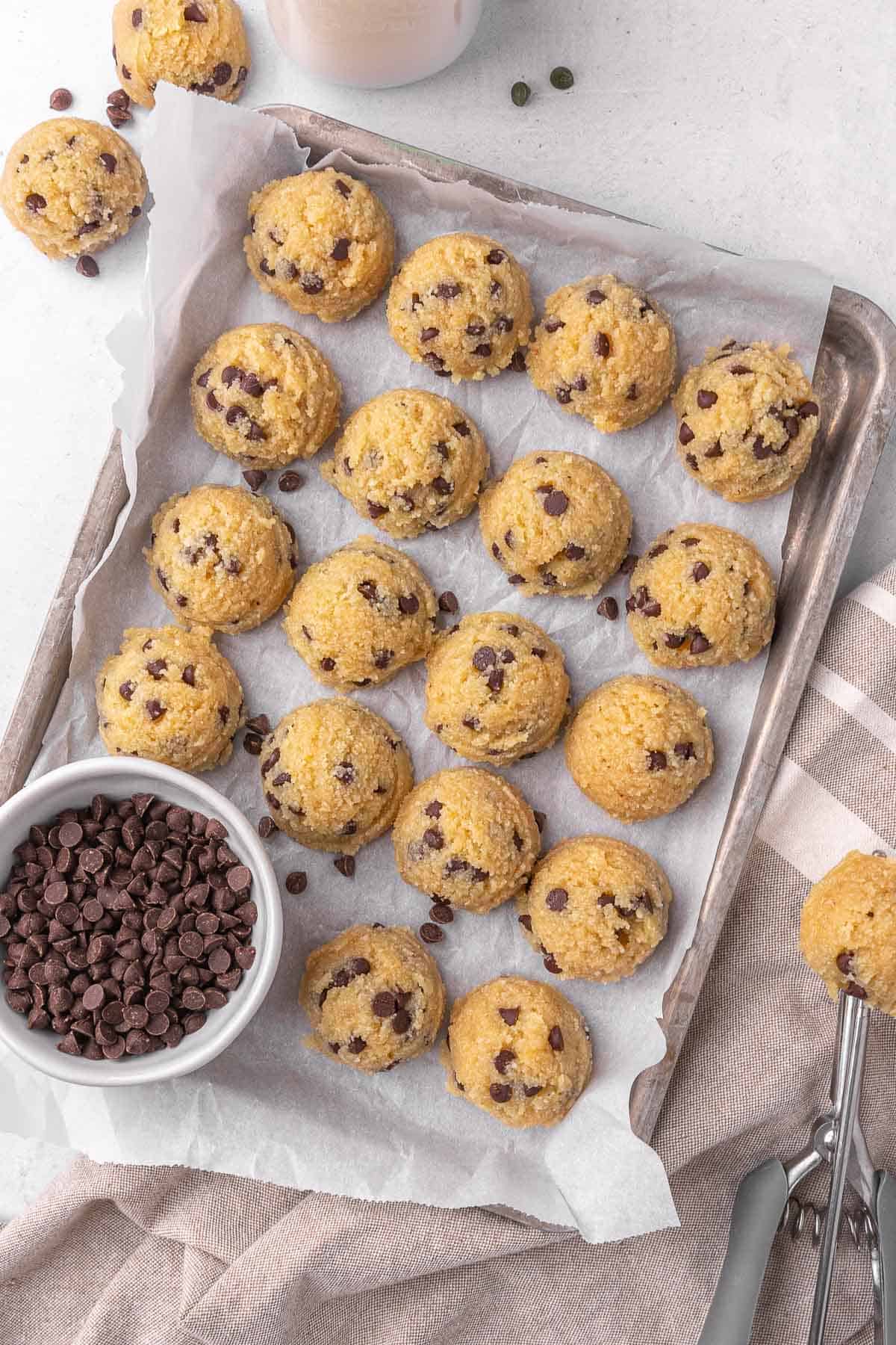 Chocolate chip edible cookie dough balls on a parchment lined sheet pan with a small dish of chocolate chips tucket inside. Edible cookie dough balls in a small high sided white bowl with more around.