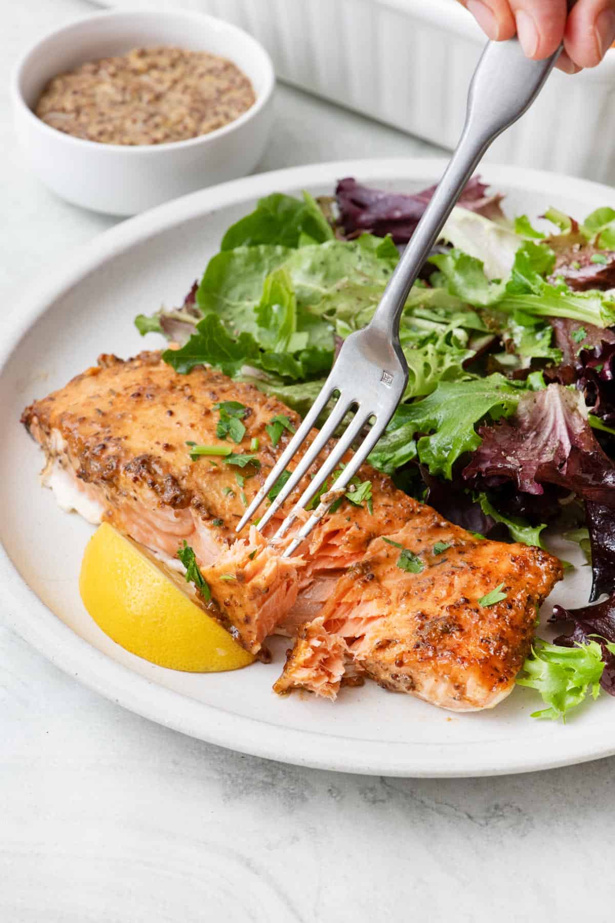 Fork flaking away a some of a honey mustard baked salmon fillet on a plat with spring mix and a small dish of honey mustard nearby.