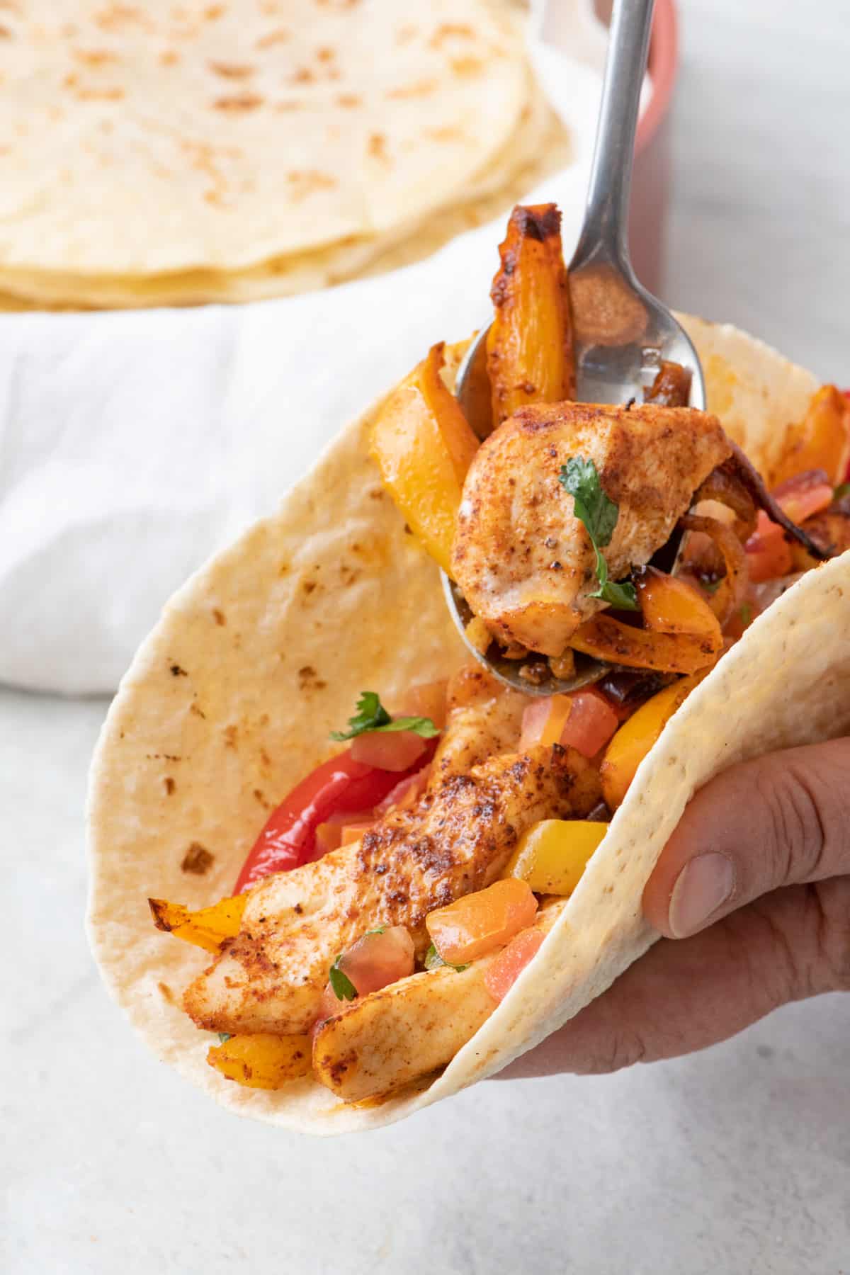 Spoon adding chicken fajita mixture into a homemade flour tortilla with extra shells in the background.