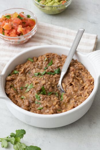 Refried beans in a serving bowl with a spoon dipped in, garnished with fresh cilantro and small bowls of salsa and guacamole nearby.