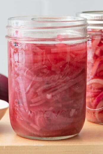 2 jars of pickled red onions, one without a lid and extra red onions nearby.