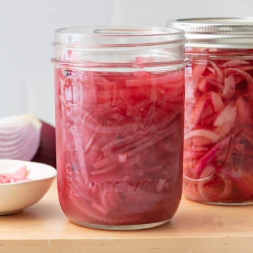 2 jars of pickled red onions, one without a lid and extra red onions nearby.