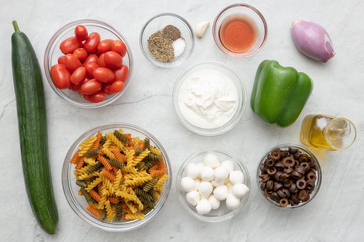 Ingredients before prepping: english cucumber, bowl of cherry tomatoes, bowl of tricolor rotini, small bowls of seasoning, vinagar, yogurt, mozzerella balls, and sliced black olives, green bell pepper, shallot, and olive oil.