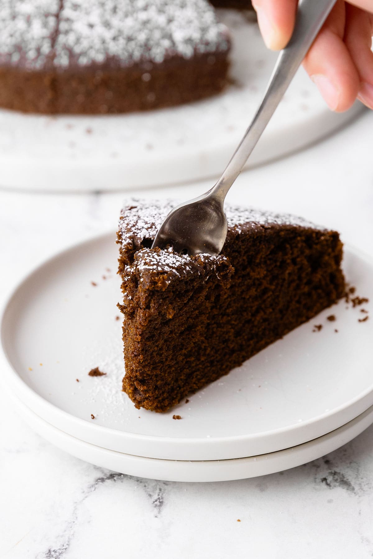 Closeup of fork digging into a slice of chocolate olive oil cake on a small white plate.