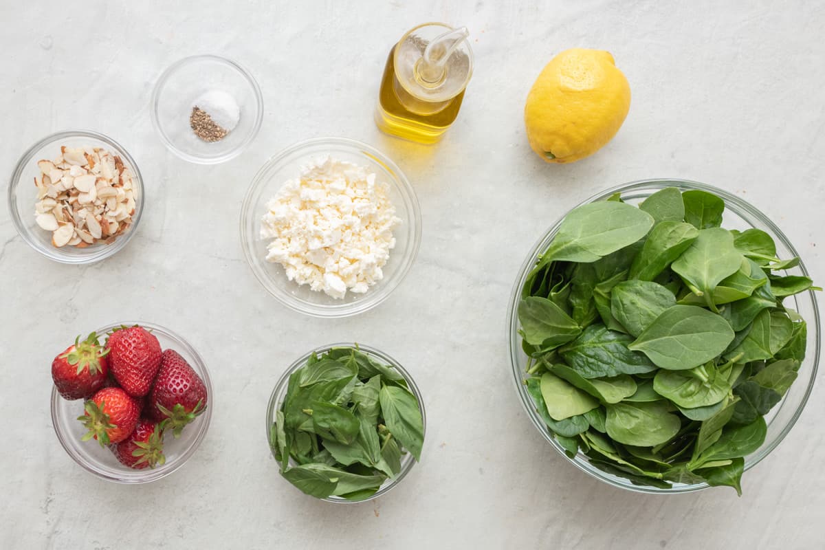 Ingredients for recipe: sliced almonds, salt and pepper, whole strawberries, crumbled feta, basil leaves, oil, lemon, and fresh spinach.