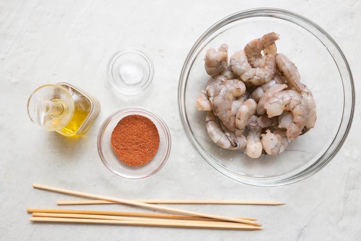 Ingredients for recipe in individual bowls and jar: oil, salt, bbq seasoning, shrimp, plus wooden skewers.