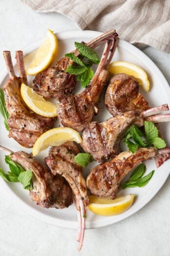 Grilled lamb chops on a round white plate garnished with mint leaves and lemon wedges.