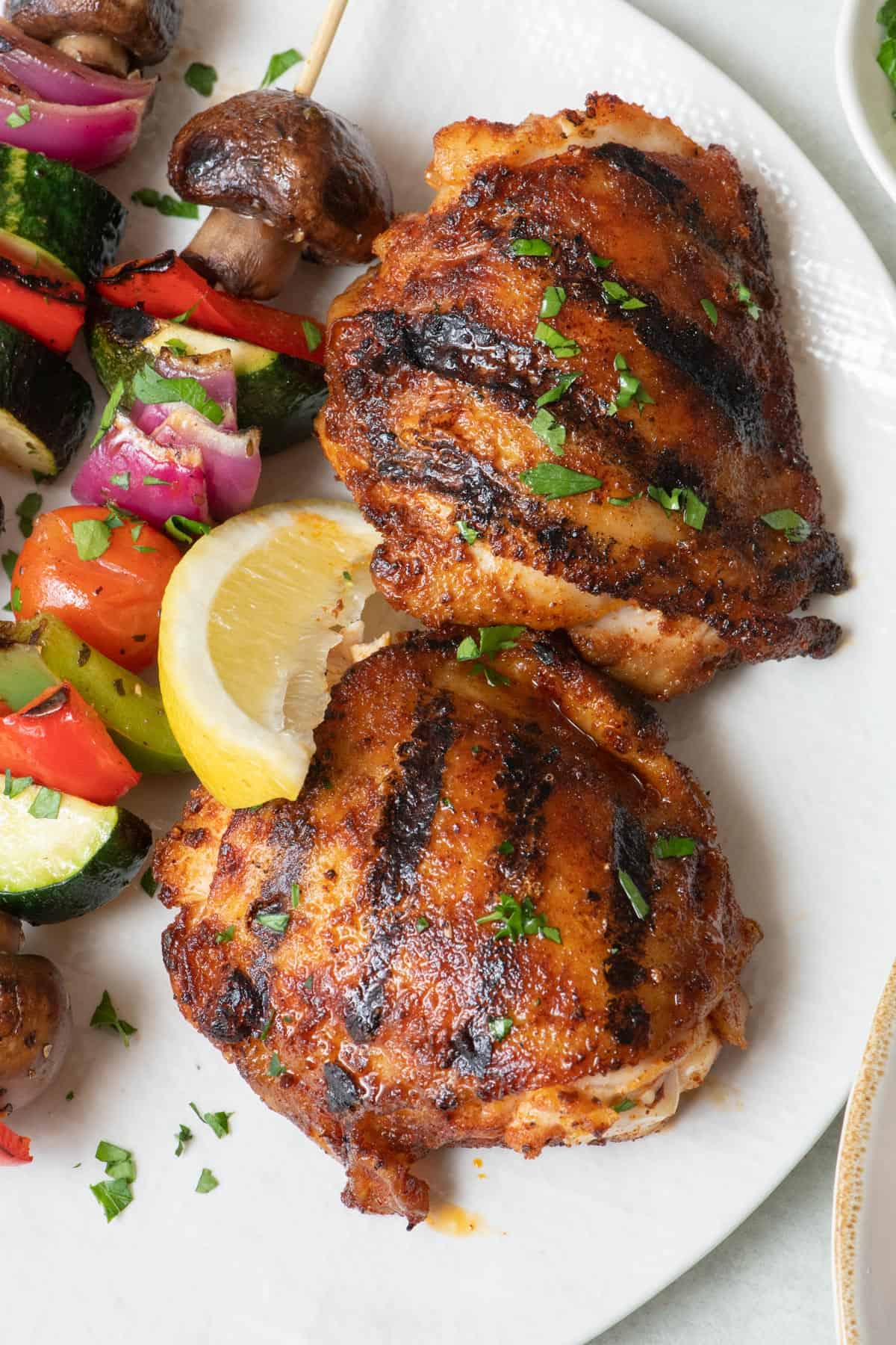 Two grilled chicken thighs on a plate next to veggie skewers, garnished with parsley and a lemon wedge.