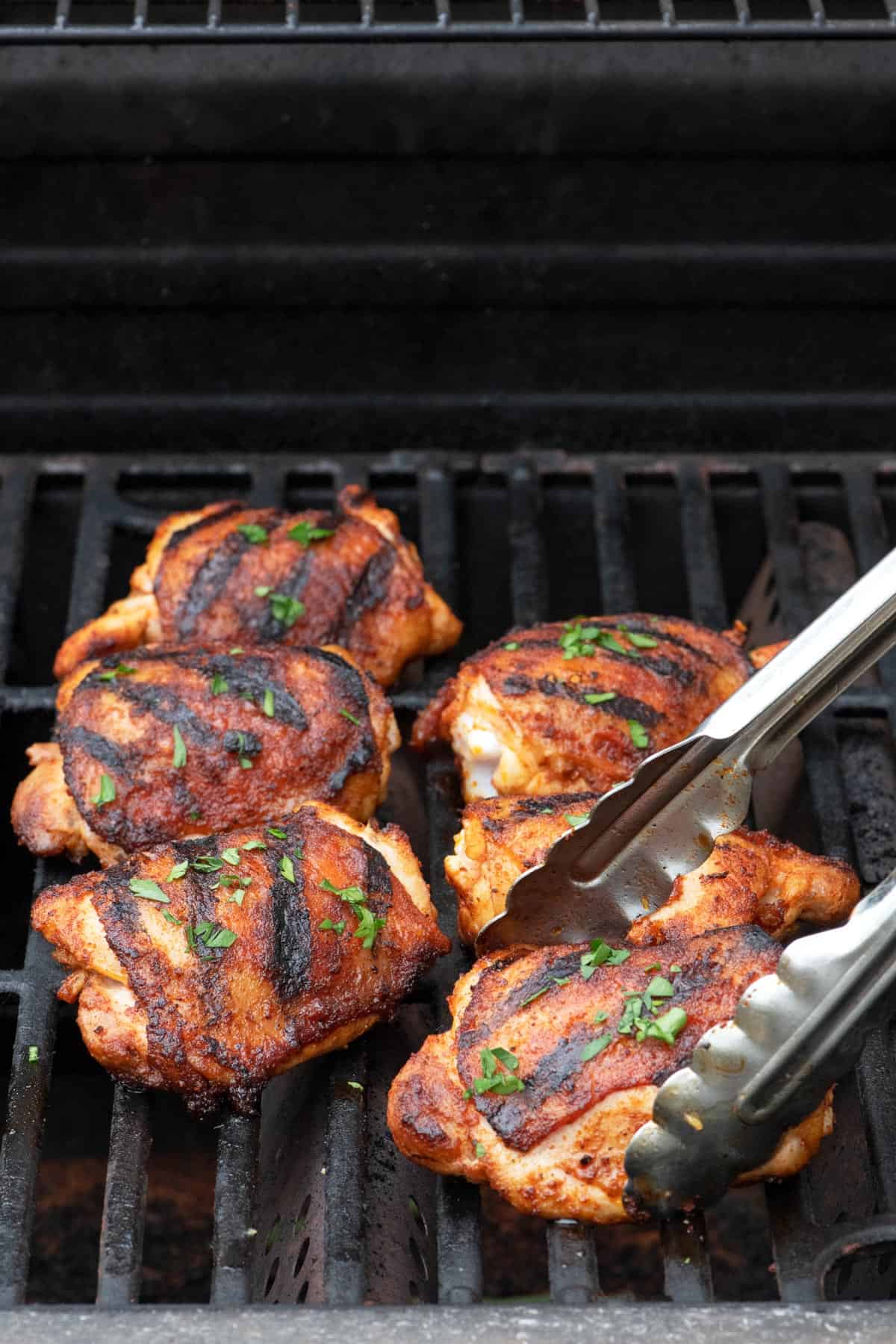 How to grill chicken thighs: 6 grilled chicken thighs on the grill with tongs flipping lifting one up.