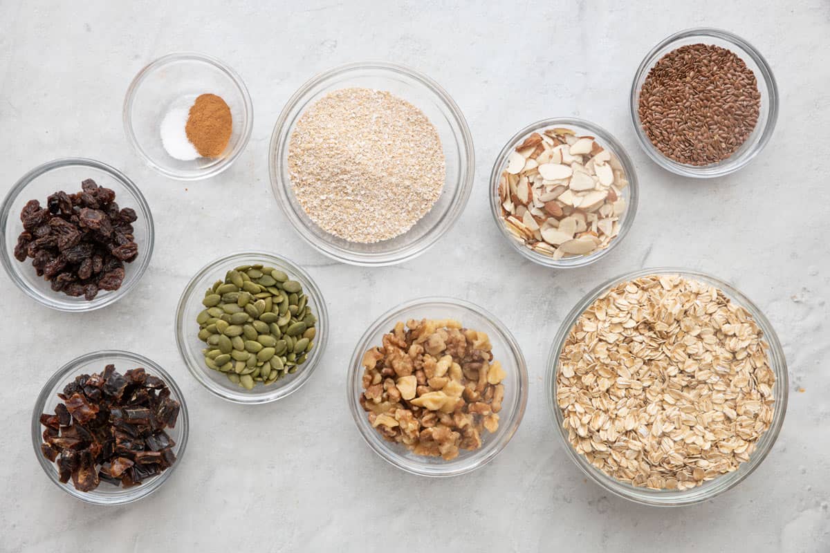 Ingredients for recipe in individual bowls: raisins, chopped dates, cinnamon and salt, pumpkin seeds, wheat bran, chopped walnuts, sliced almonds, flaxseed, and old-fashioned oats.