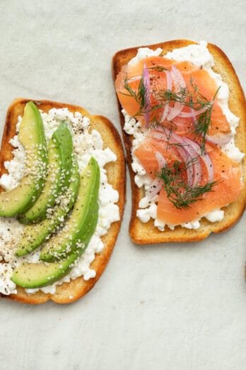 4 cottage cheese toasts with various toppings, from top to bottom: 1- sliced cucumbers and pesto, 2- smoked salmon, red onion, and fresh dill, 3- sliced avocado and sesame seeds, and 4- diced tomatoes and za'atar. Extra toppings in small bowls nearby.
