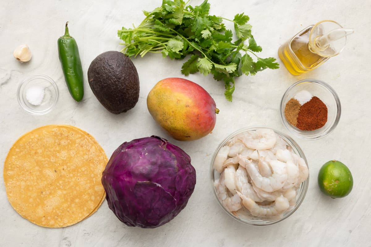 Ingredients for recipe: garlic, salt, jalapeรฑo, avocado, corn shells, head of red cabbage, mango, cilantro, oil, spices, raw shrimp, and lime.