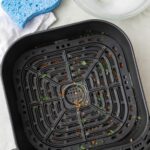 A dirty air fryer basket with cleaning supplies and soapy water nearby.