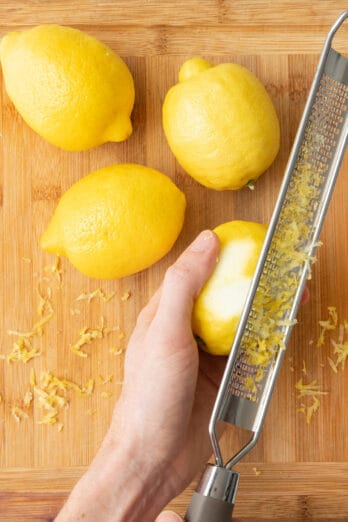 Lemons on a cutting board with a hand holding one while using a microplane to zest it.