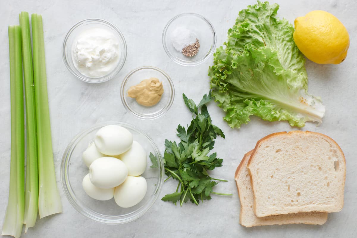 Ingredients for recipe before prepping: celery stalks, yogurt, boiled eggs, dijon mustard, salt and pepper, parsley, lettuce, lemon, and bread slices.