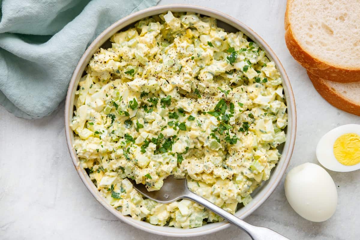Egg salad in a large shallow serving bowl with spoon dipped in, garnished with fresh parsley and black pepper.