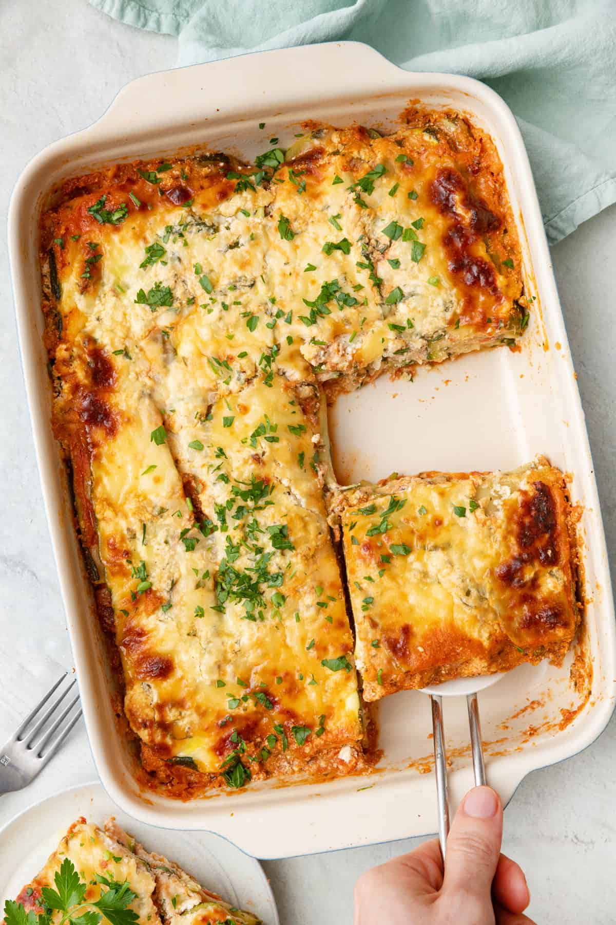 Zucchini lasagna in a baking dish with a serving removed and plated to the side and another large piece being lifted from pan