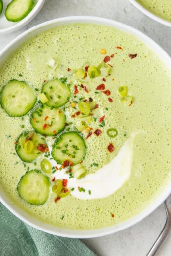 Bowl of cold cucumber gazpacho, garnished with sliced cucumbers, red pepper flakes, and green onions.