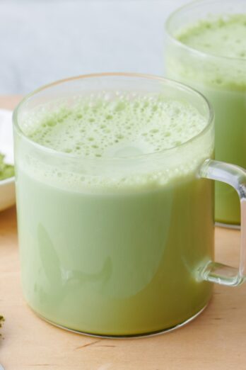 Matcha latte in a glass mug with matcha powder on a spoon and small dish.