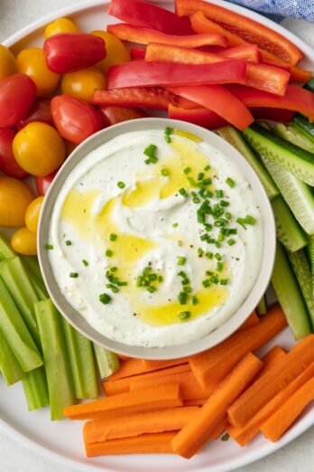 Whipped cottage cheese in a bowl garnished with chives and olive oil. Bowl on a platter surrounded by fresh veggies cut into sticks, including red bell peppers, zucchini, carrots, celery, and tomatoes.