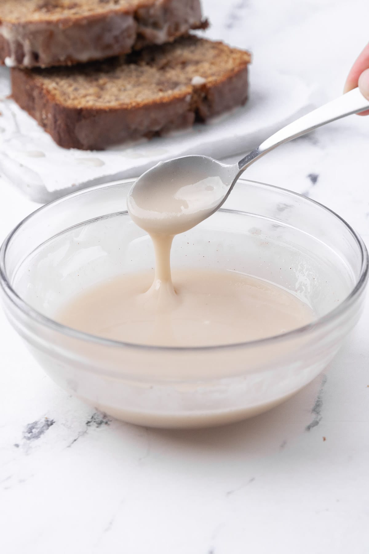 Homemade glaze in a bowl with a spoon lifting some up and a few slices of quick bread nearby.