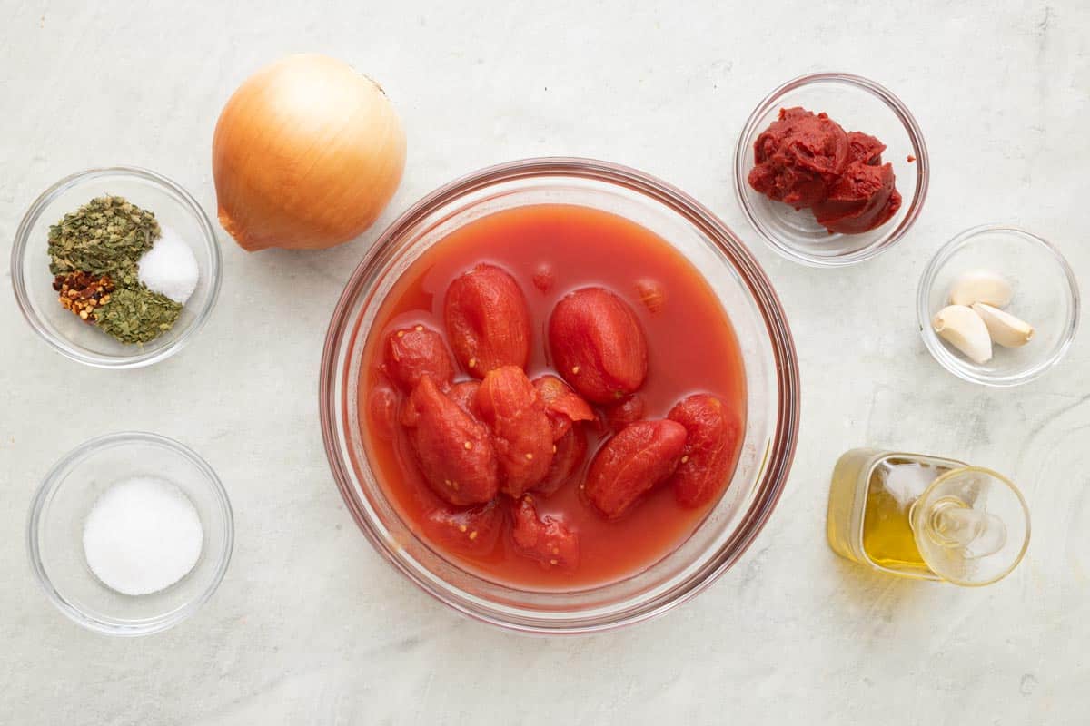 Ingredients for recipe: seasonings, salt, sugar, yellow onion, canned whole tomatoes in bowl, tomato paste, garlic cloves, oil.