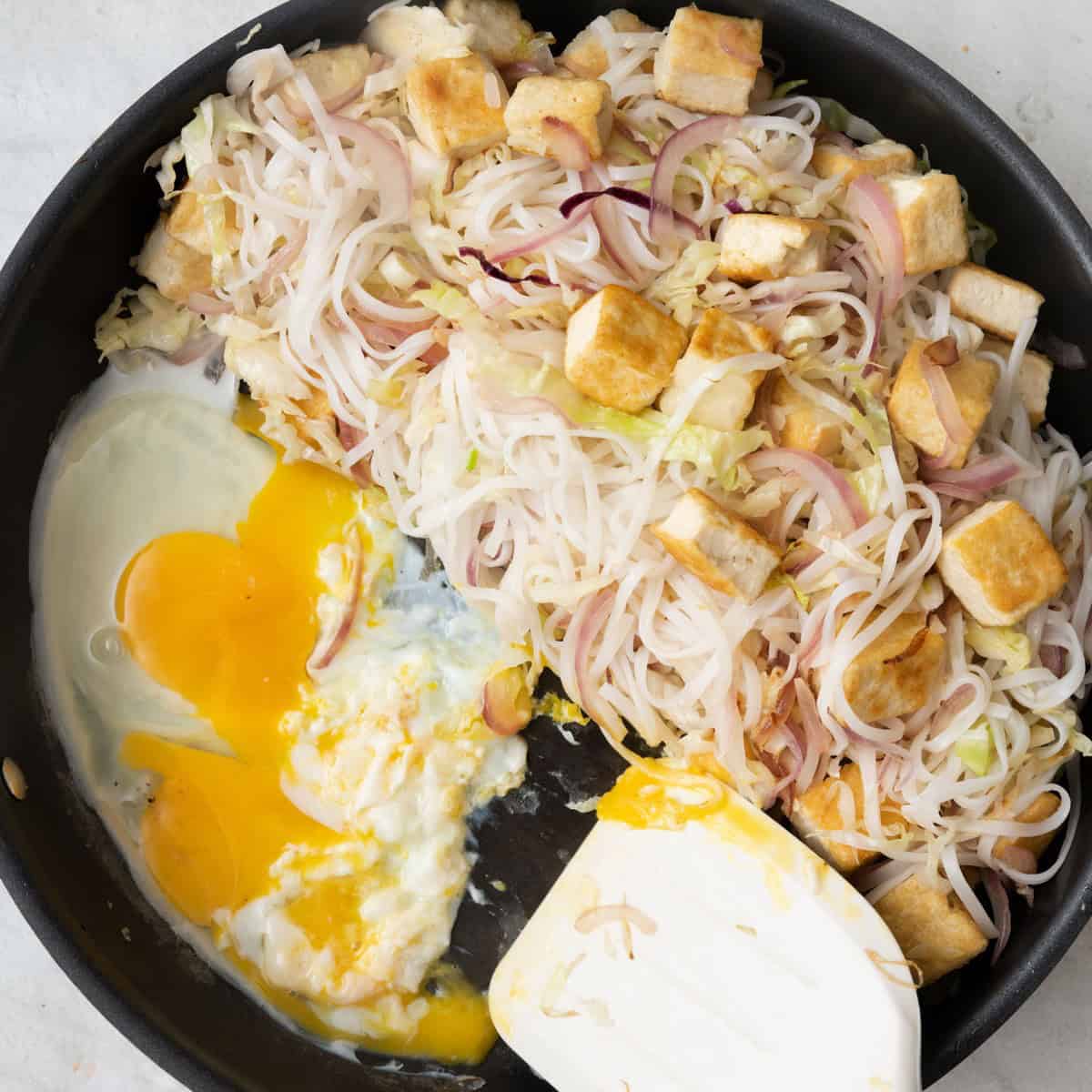 Blanched noodles and cooked tofu added and pushed to side of wok with uncooked eggs added and with spatula pushing them around to scramble.