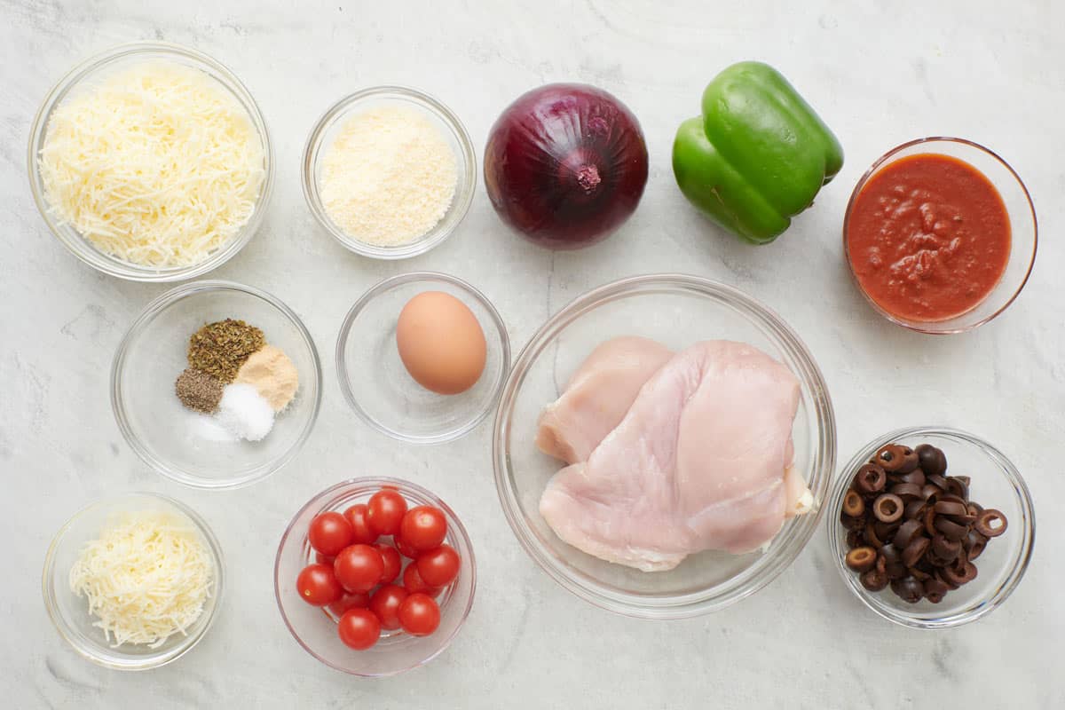 Ingredients for recipe: shredded cheeses, spices, egg, cherry tomatoes, red onion, green bell pepper, pizza sauce, raw chicken breasts, and sliced black olives.