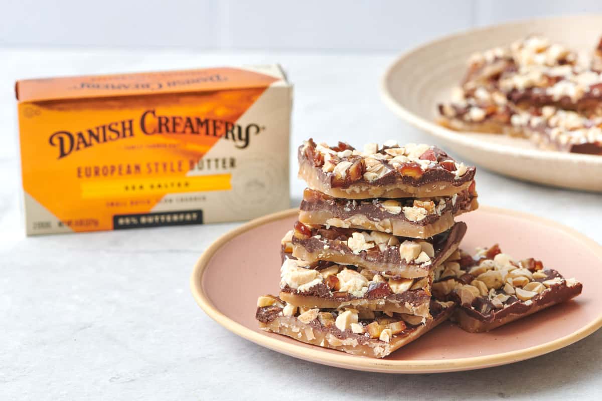 Toffee with chocolate, peanuts, and dates on top stacked on a plate next to a box of Danish Creamery Sea Salted European Style Butter.