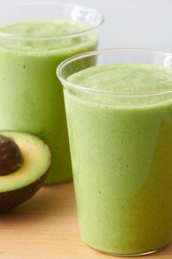 Two cups of avocado smoothie with half avocado nearby.