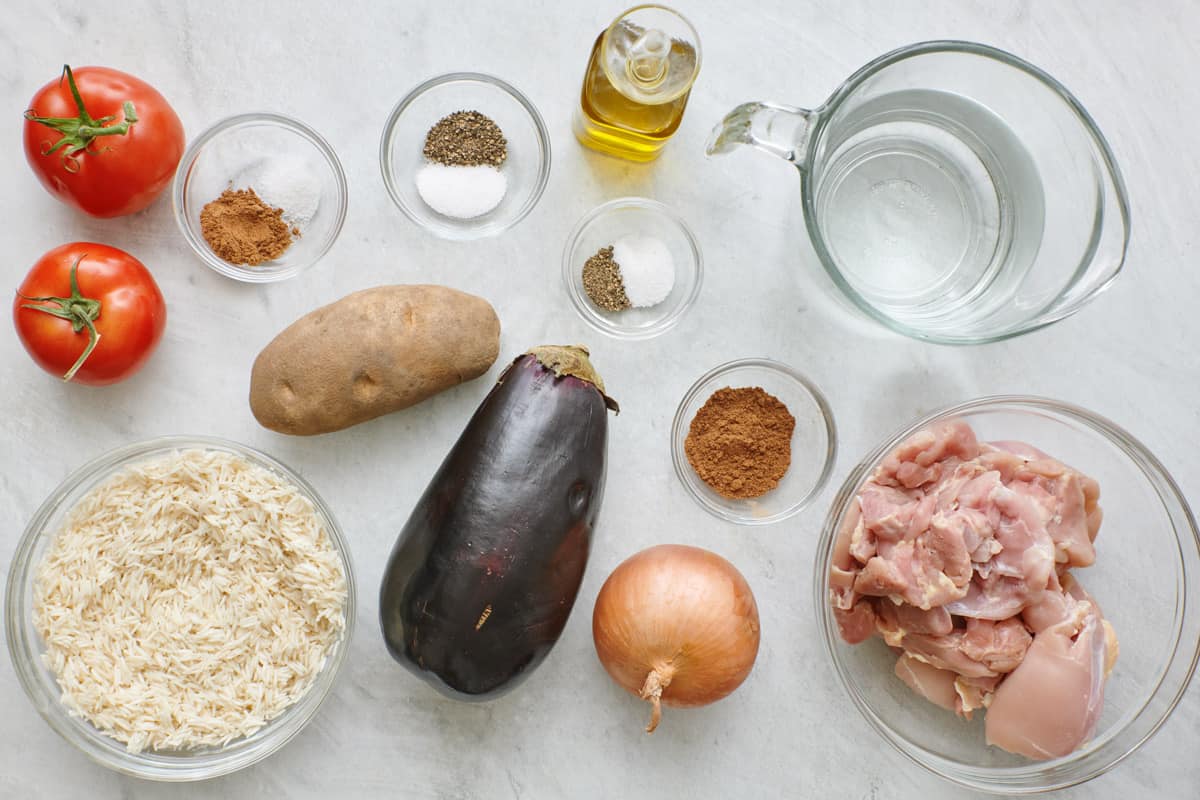 Ingredients for recipe before prepping: tomatoes, spices and seasonings, uncooked rice, potato, eggplant, onion, chicken thighs, and water.