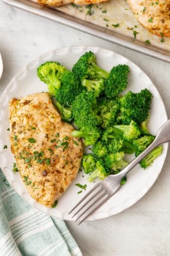 A white plate with baked ranch chicken and a side of broccoli with a tray of other baked chicken and a seafoam green and white striped cloth napkin nearby.