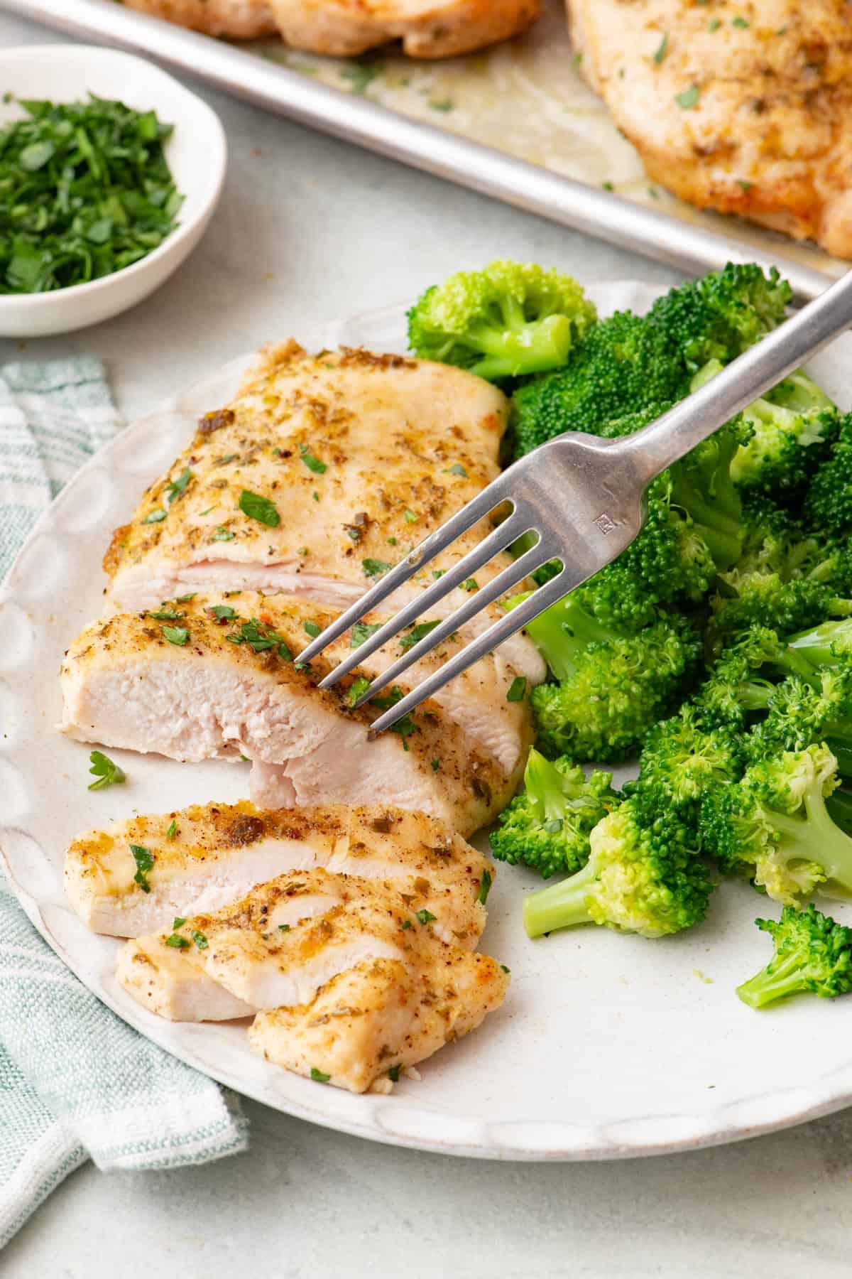 Sliced baked ranch chicken being stabbed by a fork on a white plate with a side of broccoli; a bowl of parsley and tray of more cooked chicken nearby.