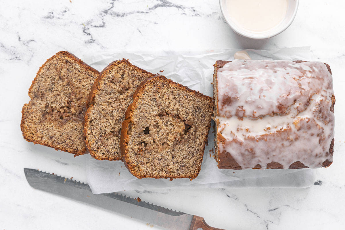Cinnamon swirl banana bread on a marble cutting board cut halfway with a few slices laid down to show cinnamon swirl inside; small dish of extra glaze nearby.