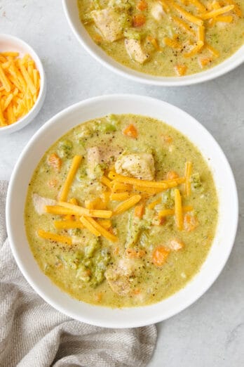 Two bowls of creamy chicken broccoli soup with shredded cheddar cheese on top.