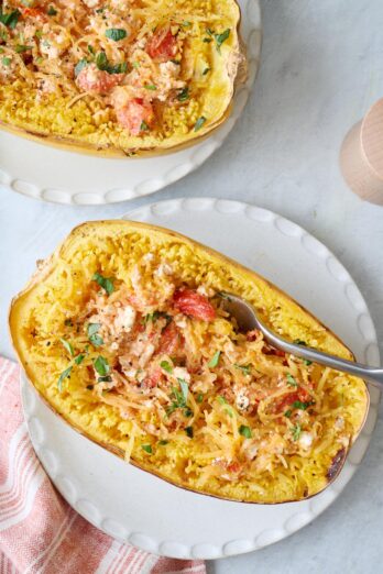 A fork scooping from a feta spaghetti squash made with cherry tomatoes and garlic; another spaghetti squash nearby.