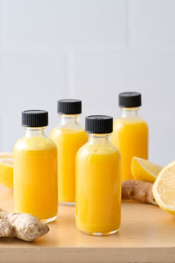 Ginger shots in small glass bottles on a wooden table with lemons and ginger roots nearby.