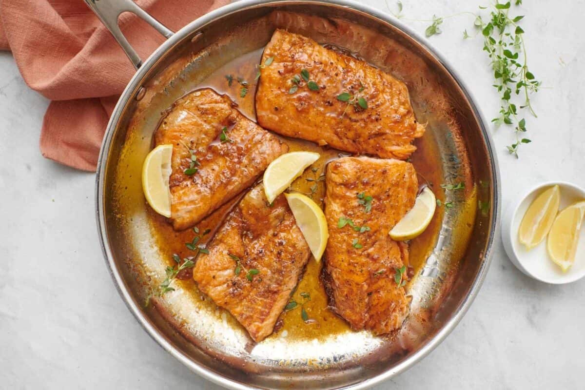 4 honey lemon salmon fillets in a skillet with sauce, garnished with fresh parsley and lemon wedges.