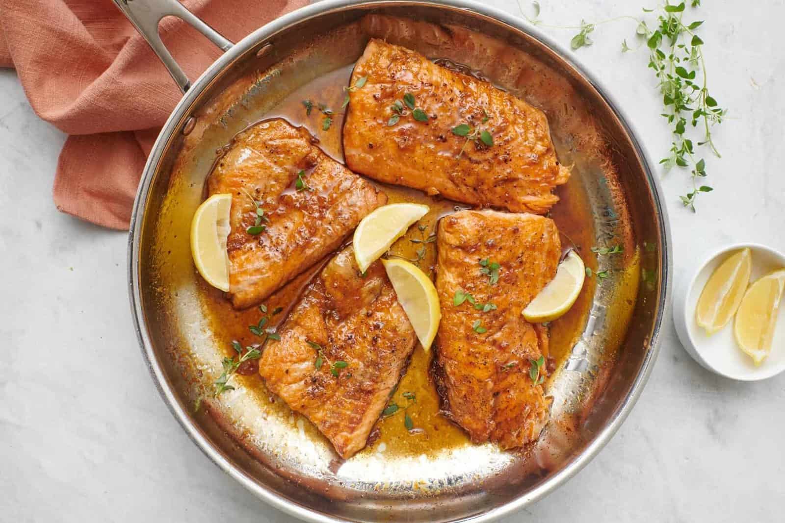 4 honey lemon salmon fillets in a skillet with sauce, garnished with fresh parsley and lemon wedges.