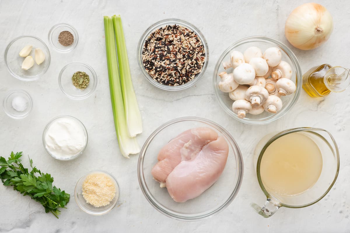 Ingredients for recipe before prepping, in individual bowls: garlic, salt, pepper, dried oregano, yogurt, parsley, celery stalks, parmesan, wild rice, chicken breasts, mushrooms, broth, onion, and oil.