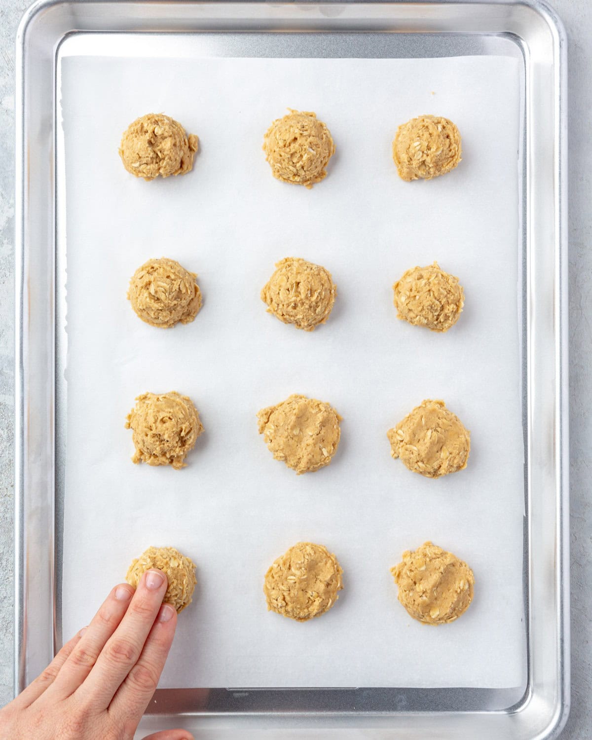 Scooped cookie dough on a parchment lined baking sheet. Hand pressing down a cookie dough ball with a few already pressed.