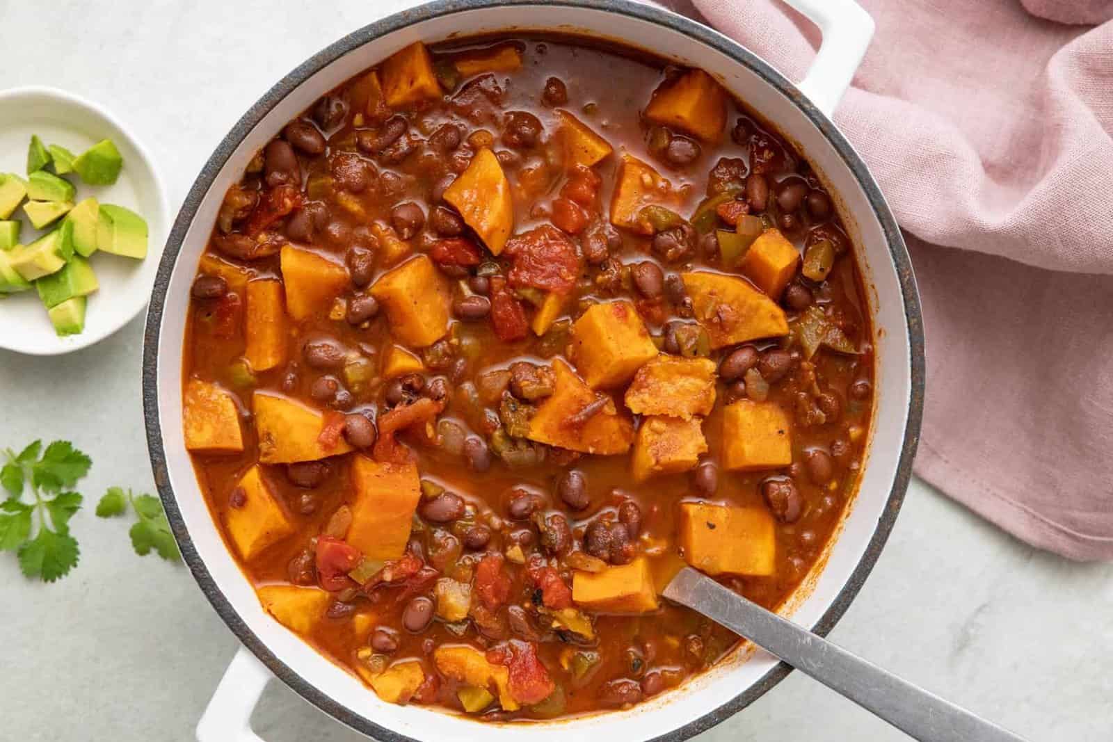 Sweet potato chili in pot with a ladle dipped inside. Small bowl of diced avocado nearby.