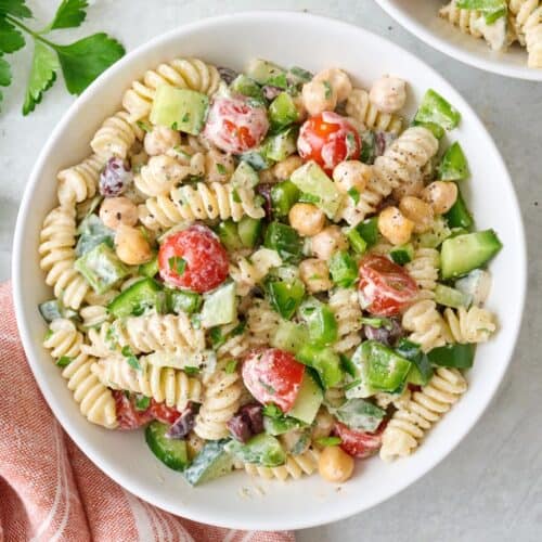 Closeup of a small bowl of tahini pasta salad with tomatoes, cucumbers, chickpeas, bell pepper, parsley, and olives.