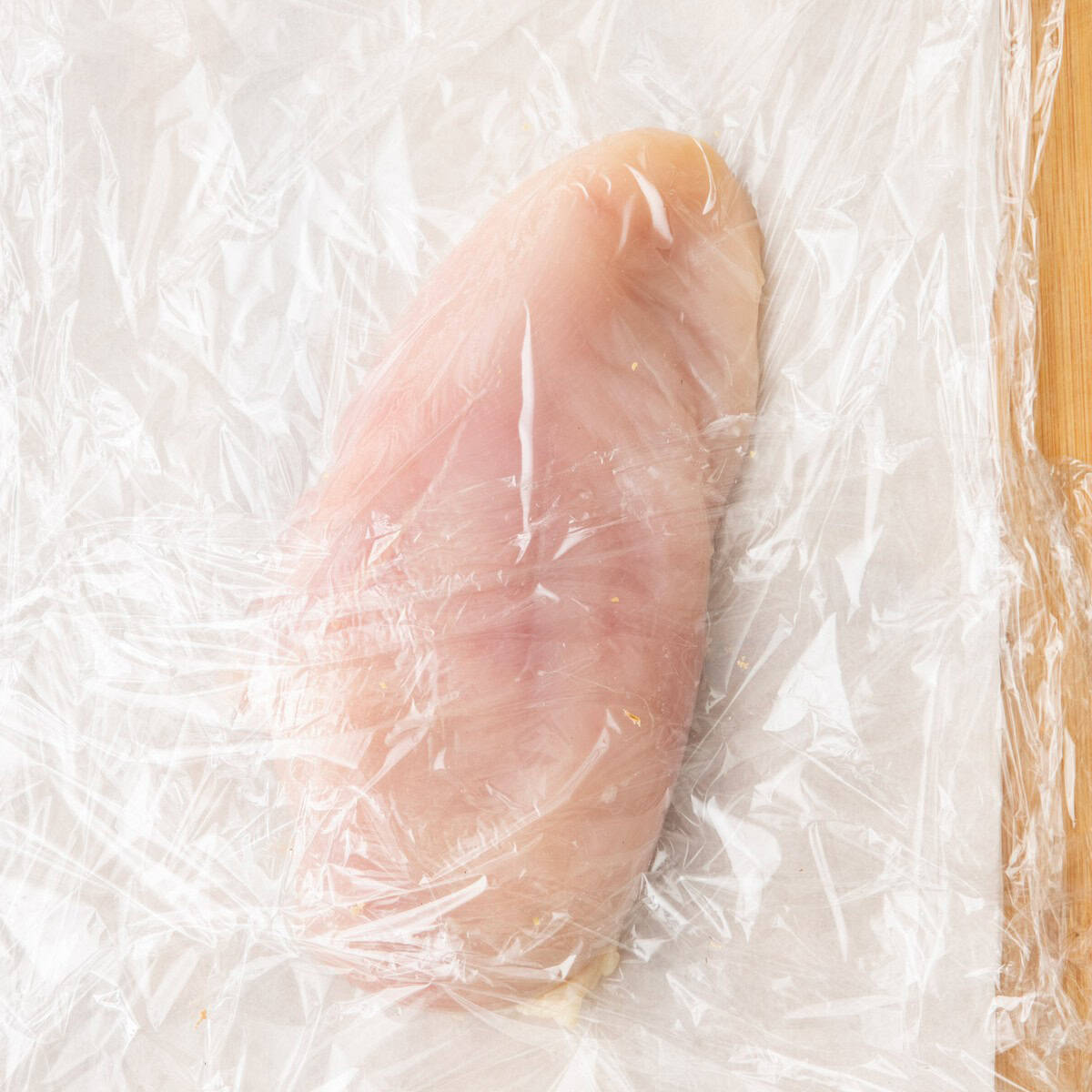 Chicken breast on a sheet of parchment paper covered with plastic wrap.
